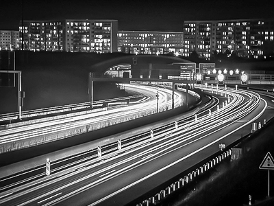 Künstlerisches Schwarz-Weiß-Foto der Autobahn in Jena bei Nacht, ein Werk von Fotograf Michael Schalansky. Es zeigt die fließenden Lichtspuren des Verkehrs, die im Kontrast zu den feststehenden Gebäuden im Hintergrund stehen und eine tiefgründige Atmosphäre schaffen.